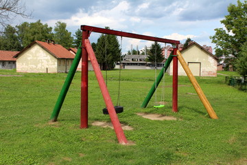 Vintage retro colorful wooden outdoor public playground equipment in shape of swing with classic plastic and rubber tyre seats surrounded with grass in front of destroyed buildings of abandoned