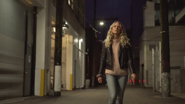 Slow motion of young blonde woman in jeans and leather jacket posing and walking on streets in downtown after sunset in the evening