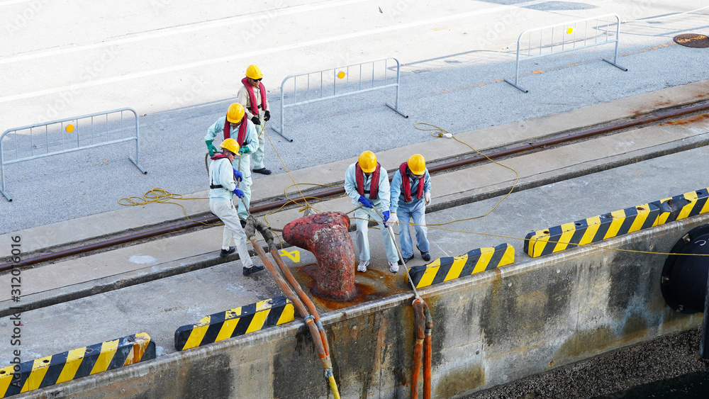 Wall mural a group of japanese port workers in uniform and orange helmets fasten ship mooring ropes to the boll