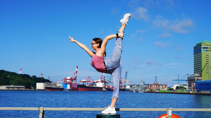 very flexible young girl gymnast does the splits on the background of cargo cranes at an industrial port and blue ocean water. acrobat in life style clothes on a sunny day at the observation deck