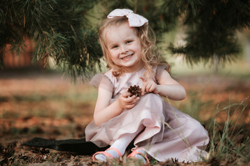 little smiling girl with a bow on her head and in a dress with sits under a tree. Child enjoys playing with cones