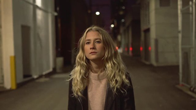 Slow motion of young blonde woman in jeans and leather jacket posing and walking on streets in downtown after sunset in the evening