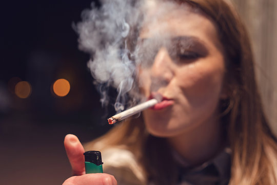 Close up portrait of young caucasian woman on the street at night lighting cigarette with lighter smoking in the city outdoor smoke