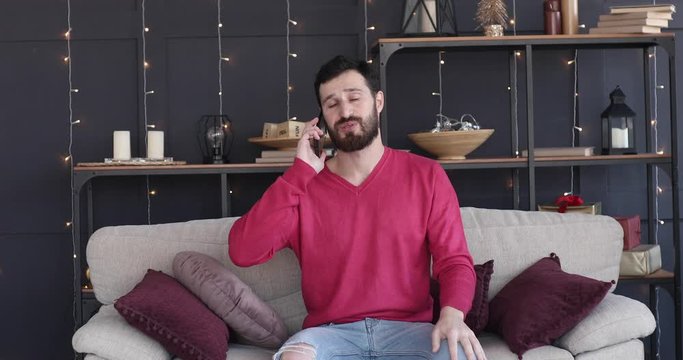 Happy young bearded man uses smartphone while sitting on a sofa at home. Man browses  internet, watches videos and uses social networks at home.