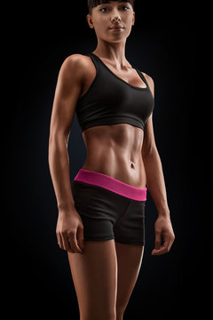 Beautiful sexy female slim tanned body. Cheerful attractive young fitness woman in black top and black shorts isolated over black background