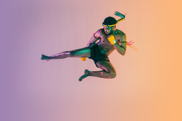 Naklejka na ściany i meble Muay thai. Young man exercising thai boxing on gradient background in neon light. Fighter practicing, training in martial arts in action, motion. Healthy lifestyle, sport, asian culture concept.