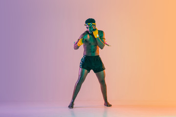 Muay thai. Young man exercising thai boxing on gradient background in neon light. Fighter practicing, training in martial arts in action, motion. Healthy lifestyle, sport, asian culture concept.
