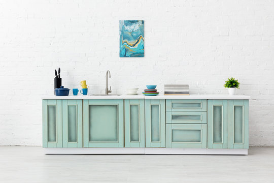 White And Turquoise Kitchen Interior With Sink, Tableware And Abstract Painting On Brick Wall