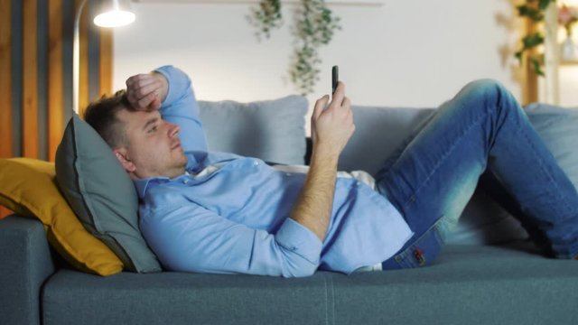 Man using smartphone for watching video or chatting in social media. Man seat on sofa in modern living room. Young boy relaxing at home in evening.