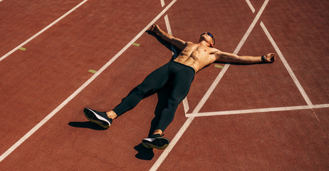 Overhead view image of young handsome shirtless athlete man lying on racetrack at stadium taking...