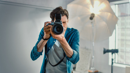 In the Photo Studio with Professional Equipment: Portrait of the Famous Photographer Holding State of the Art Camera Taking Pictures with Softboxes Flashing in Background. - obrazy, fototapety, plakaty