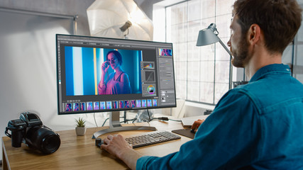 Professional Photographer Sitting at His Desk Uses Desktop Computer in a Photo Studio Retouches. After Photoshoot He Retouches Photographs of Beautiful Female Model in an Image Editing Software - obrazy, fototapety, plakaty