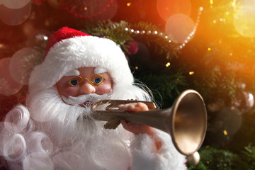 Close-up of a Santa Claus doll  with white beard, red coat and trumpet in front of a Christmas tree.