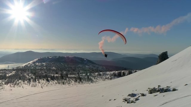 A paraglider flies on a bright parachute with a red plume of smoke against the background of a winter mountain valley. bright paraglider in flight. active recreation and paragliding, skydiving