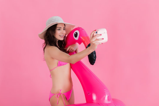 The girl in a swimsuit and hat holds a flamingo and takes a selfie a photo on the instant camera