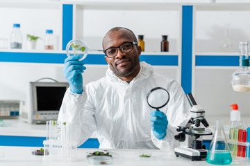 smiling african american biologist holding magnifying glass and leaves