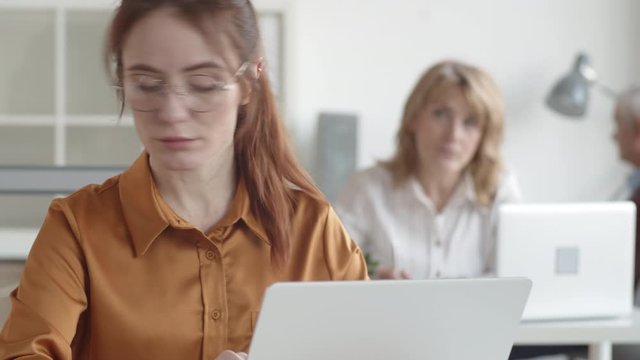 Tilting close-up shot of young Caucasian woman sitting in office with anxious face, looking at documents and typing on laptop, and middle-aged supervisor watching and fidgeting with pen in background