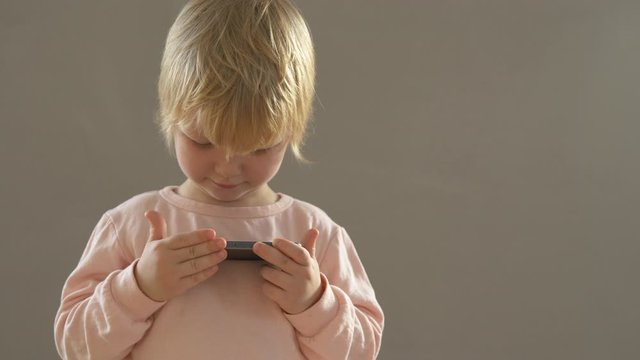 Cute blond boy in pink pajamas plays with smartphone touching touch screen with finger. child wants to quickly become adult and learn everything. Home teaching concept for lagging children