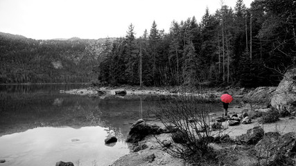 Walk around the Eibsea (Eibsee) near Zugspitze (highest mountain in Germany). Clear weather, no wind. Sea surrounded with green fir trees, grey stones and rock fragments. Wanderer with red umbrella.