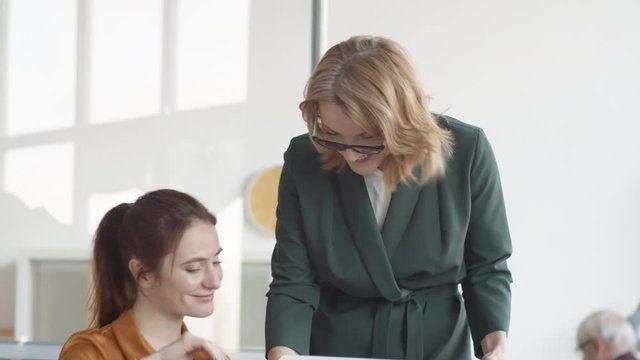 Medium shot of young Caucasian woman working on laptop in office, and middle-aged female manager coming over, showing her folder with documents and giving task