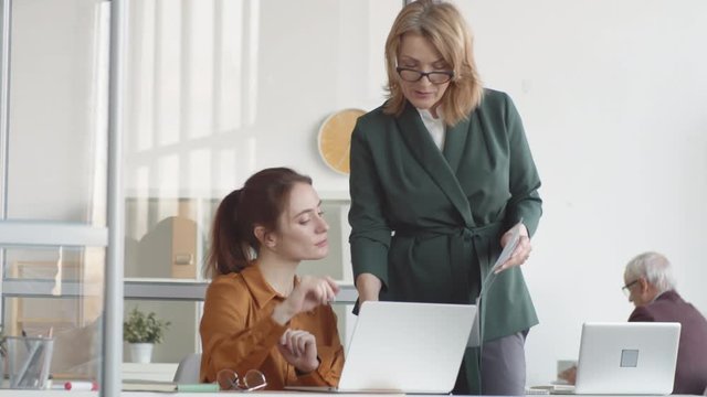 Medium shot of middle-aged female manager walking over to young secretary, showing her document in folder, asking question, woman looking up information on laptop and demonstrating on screen