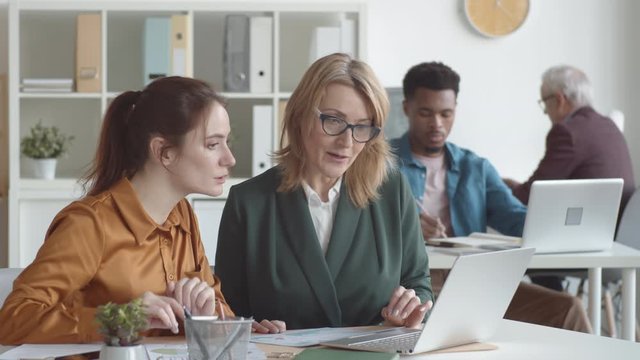 Waist-up shot of new Caucasian female employee sitting at desk together with senior mentor, who is pointing at laptop screen, smiling and explaining her work duties while girl is asking questions