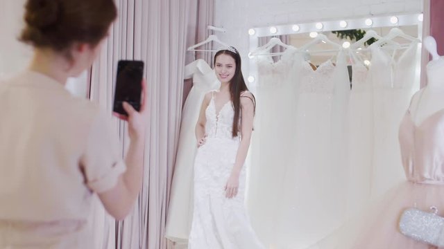 Rear view of young woman making photos of her female friend trying on trendy wedding dress using phone camera