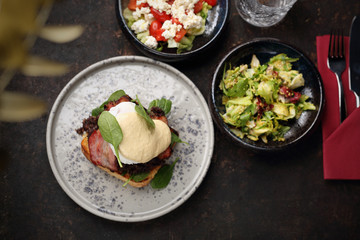 Toast with black pudding, poached egg, bacon and mayonnaise sauce served with a patch of fresh green vegetables. Food, ready dish on a dark background. Suggestion of serving the dish.Top view