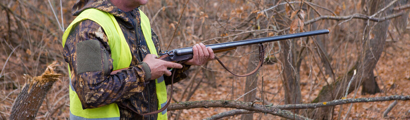 A man with a gun in his hands and an orange vest on a pheasant hunt in a wooded area in cloudy weather. Hunter with dogs in search of game.