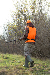 A man with a gun in his hands and an orange vest on a pheasant hunt in a wooded area in cloudy weather. Hunter with dogs in search of game.
