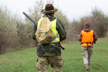 A mans with a gun in his hands and an orange vest on a pheasant hunt in a wooded area in cloudy weather. Hunters with dogs in search of game.