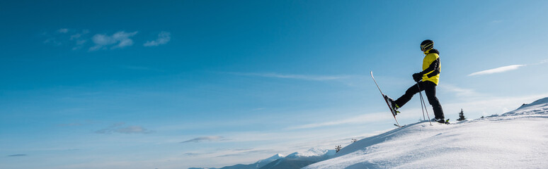 Panoramic shot of skier holding ski sticks and making step against blue sky in mountains