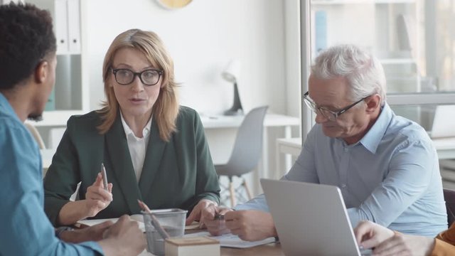 Waist-up panning shot of confident senior Caucasian business lady sitting around table in office with colleagues and talking convincingly while young woman asking question and typing on laptop