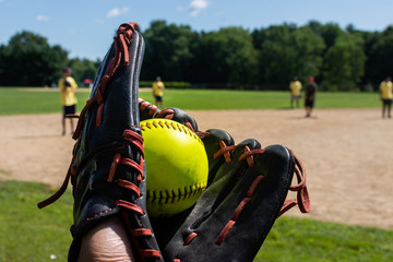 a man holds a freshly caught green softball in a black baseball glove during a softball tournament game with teammates in the background