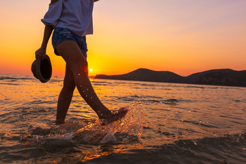 summer and travel, landscape by the sea and sunset. a girl in a white t-shirt with long hairs and hold hat. kicked the water splash .