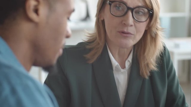 Close-up tilting shot of senior Caucasian female HR manager in glasses and jacket having serious conversation with junior black employee during appraisal meeting