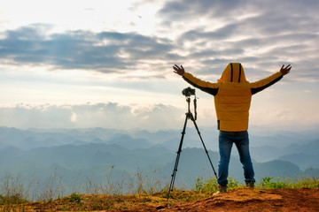 Soft blur image of the photographer with yellow jacket stand near to his camera and stand in front cliff, cloudy and show action of happy.