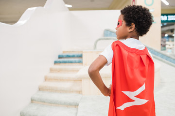 Back view of little African boy in costume of superman standing on play area