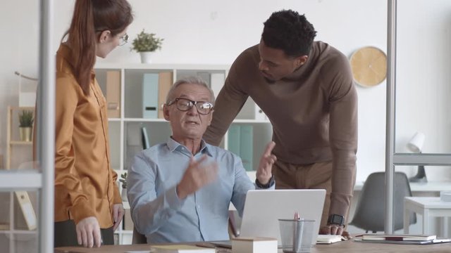 Medium shot of young black man and Caucasian woman approaching middle-aged male colleague, sitting at desk and working on laptop, and man talking to them about difficulties with project