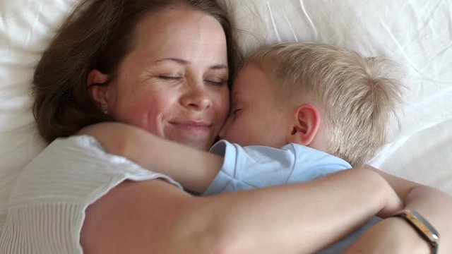Portrait of tender mom with little son in bed in the morning, they woke up and hugging. Mom hugs her beloved son lying in bed. Slow motion.