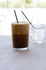 Cold frappe coffee with ice cubes on glass with black straw served on the table in caffeteria. Ice coffee in a tall glass with a lot of foam as cold summer drink.