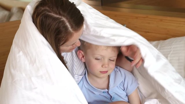 Mother hugs her little sad son on the bed under the blanket. Portrait of a sad baby on the bed, mom hugs and calms the child. Slow motion.
