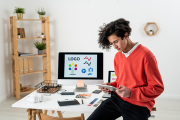 Young web designer using touchpad while sitting on desk with working supplies