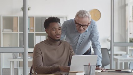 Medium shot of middle-aged Caucasian male manager approaching Afro-American employee sitting and working on laptop at desk, looking at work on screen, expressing strong opinion and surprise