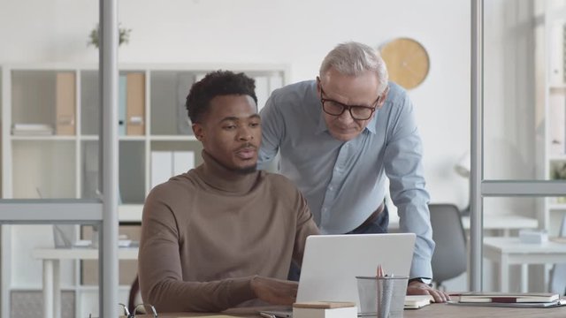 Medium shot of middle-aged Caucasian male manager approaching Afro-American employee sitting and working on laptop at desk, looking at work on screen, expressing strong opinion and surprise