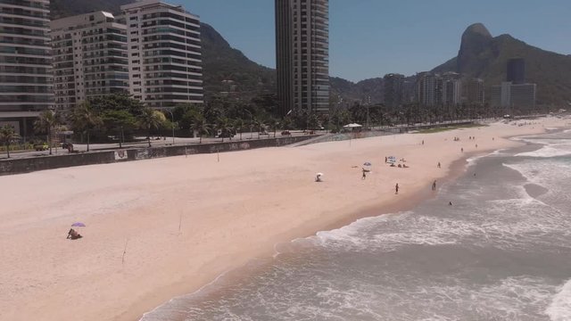 Rapid aerial approach of São Conrado beach and boulevard with high rise buildings while the waves coming ashore, Two Brothers mountain in the background against a blue sky with few people