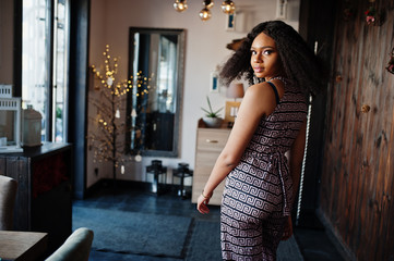 Сharming elegant young african american woman with long curly hair wearing jumpsuit posing at cafe indoor.