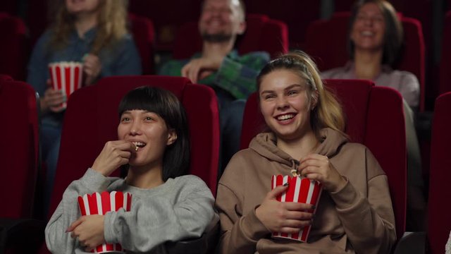 Tracking left medium shot of young people enjoying watching comedy movie sitting on comfortable seats in cinema. Diverse girls eating popcorn and laughing