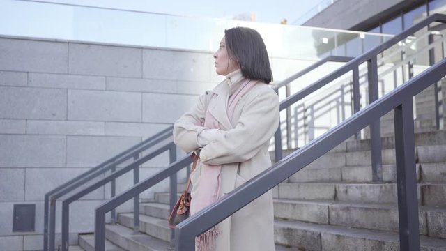 Side view of adult brunette Caucasian woman waiting for someone outdoors. Confident serious businesslady standing on stairs dressed in elegant coat. Meetings, business, lifestyle.