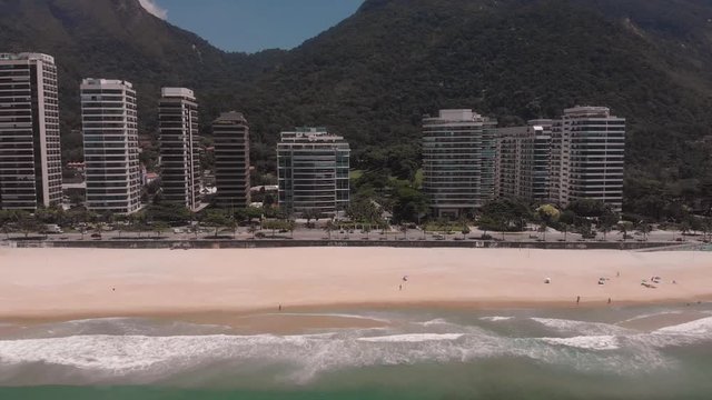 Aerial approach of São Conrado high rise buildings at the boulevard while the waves coming ashore, Two Brothers mountain in the background against a blue sky with few people
